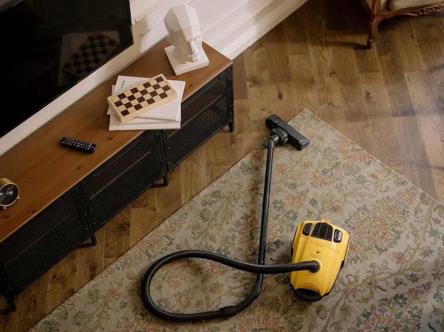 A yellow canister vacuum cleaner with a black hose and attachment is positioned on a patterned rug in a living room. The room features a wooden floor, a black metal and wood media console against a wh