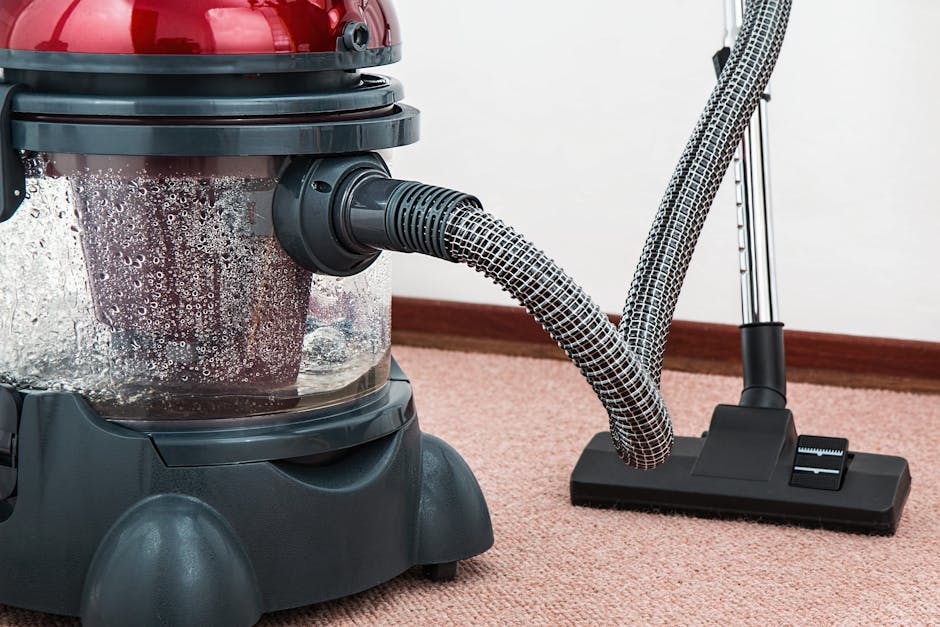 A yellow canister vacuum cleaner with a black hose and attachment is positioned on a patterned rug in a living room. The room features a wooden floor, a black metal and wood media console against a wh
