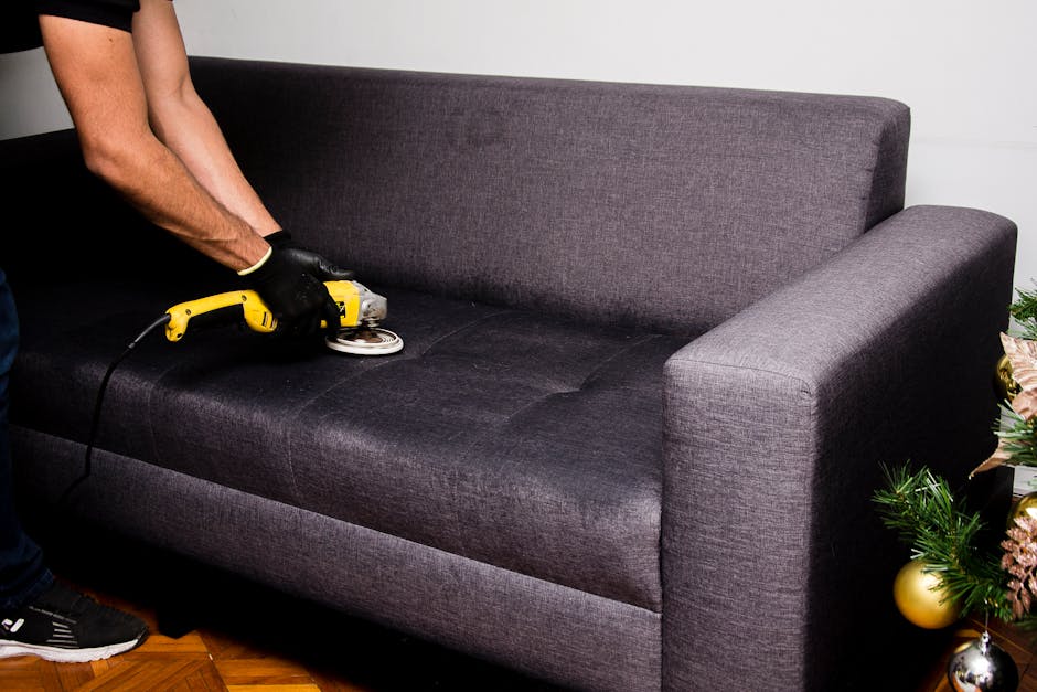 A person wearing a dark jacket is vacuuming a square decorative cushion with a yellow and white geometric pattern, placed on a dark fabric sofa in a well-lit room, demonstrating surface cleaning and m